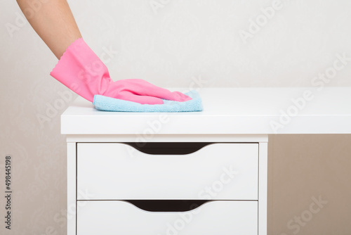 Photography Young woman hand in pink rubber protective glove holding blue dry rag and wiping dust from white work table with drawers at home office