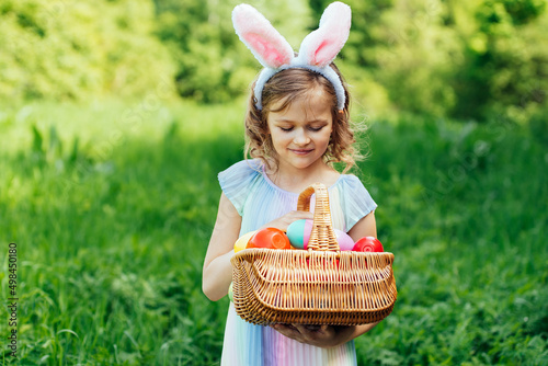 Easter egg hunt. Girl child Wearing Bunny Ears Running To Pick Up Egg In Garden. Easter tradition. Baby with basket full of colorful eggs. Bunny in basket