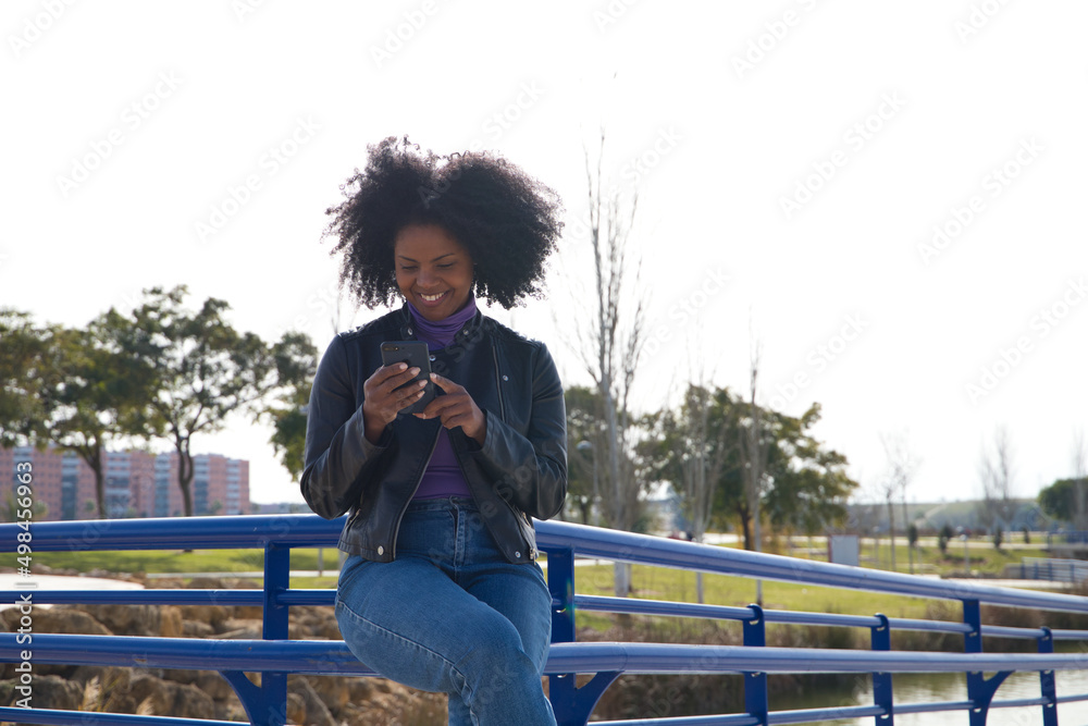 Obraz premium Young, beautiful Afro-American woman with mobile phone shopping online, checking her email and social networks in the park. The woman is wearing casual and comfortable clothes and is happy.