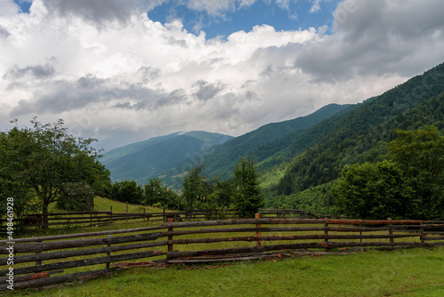Empty pasture in the mountains fenced with a wooden fence, against the backdrop of the forest, sky and clouds