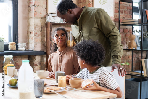 Leinwand Poster Senior African American man bending over his grandchildren at breakfast by table