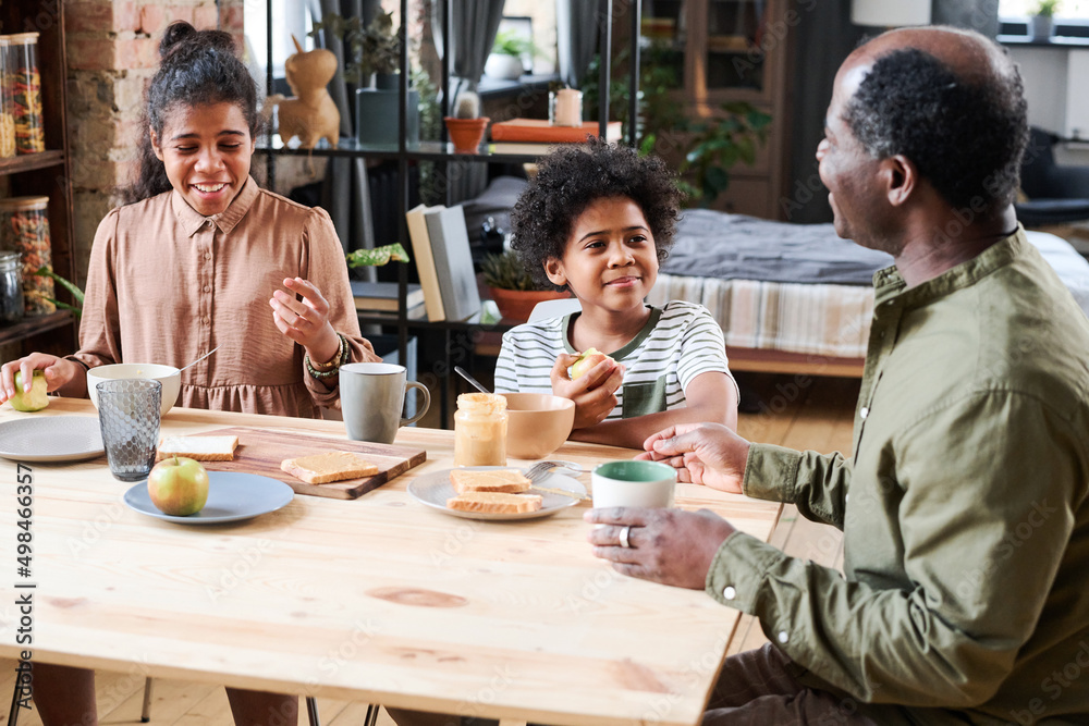 Happy cute multi-ethnic schoolkids communicating with grandfather by breakfast while sitting by served table and having snack