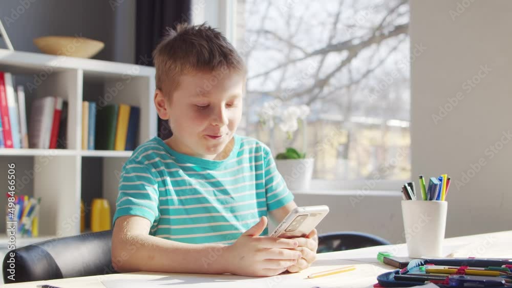 Boy is Doing his Homework at the Table. Cute Child is Learning at Home with the Help of Smartphone device and School Education Textbooks. Study and Entertainment Concept . Boy is Doing his Homework at the Table. Cute Child is Learning at Home with the Help of Smartphone device and School Education Textbooks. Study and Entertainment Concept .