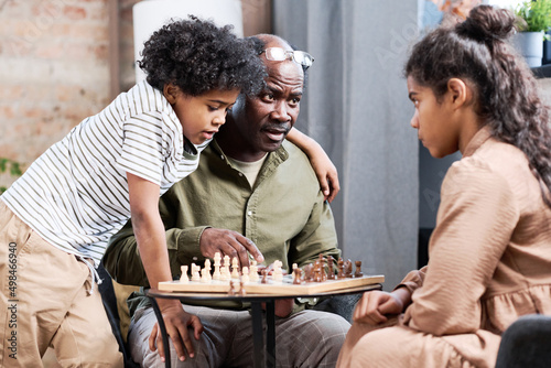 Senior African American man and his two grandchildren playing chess together while sitting by small table with figures on board