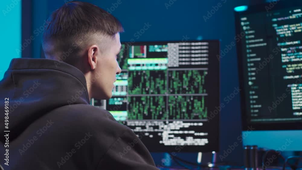 Young hacker at work close-up. The man looks at two monitors in turn, on which the implementation of the code and the lines of the program. The programmer is wearing a black sweatshirt