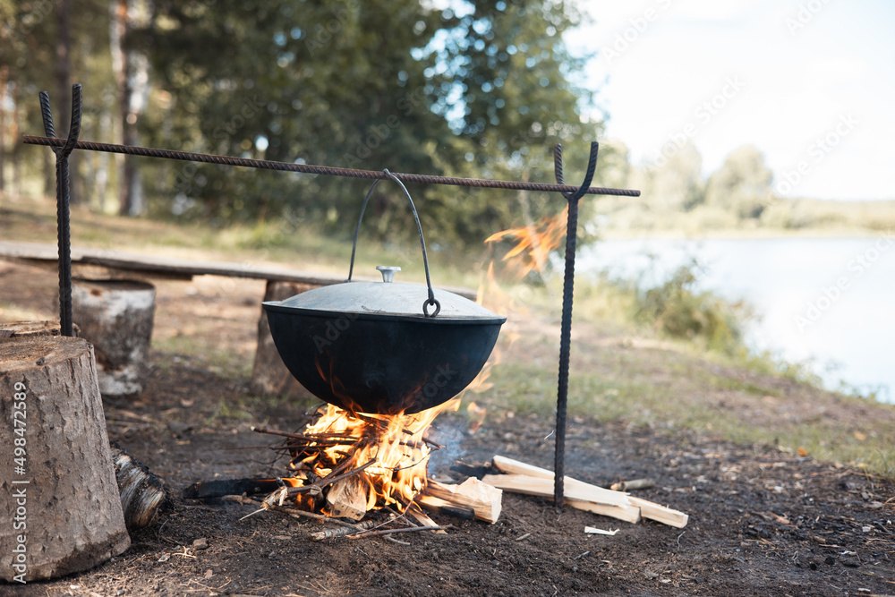Cooking in cauldron on open fire in nature. Bowler on bonfire in forest. Pot from cast iron on campfire on background of river. Copy space. Cooking in cauldron on open fire in nature. Bowler on bonfire in forest. Pot from cast iron on campfire on background of river. Copy space.