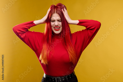 Young woman with red hair holding head by hands on yellow background
