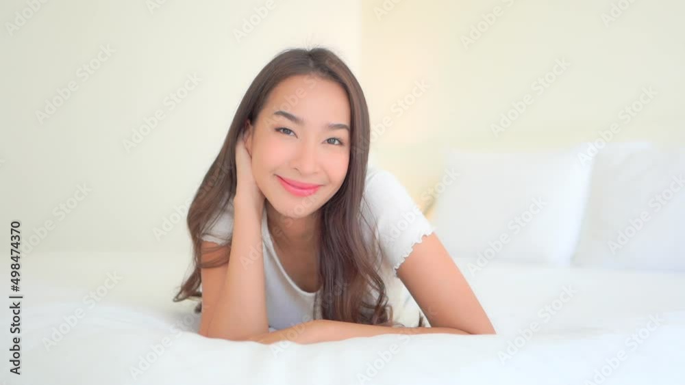Attractive Asian woman in white pajama top lying on large soft bed with white linen. Smiling happy lady in bedroom smiles and looks into camera playful daydream daytime.