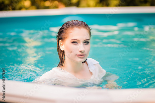 Portrait of a beautiful girl with wet hair floating in the pool, wearing a white shirt. Rest in the water in hot weather. 