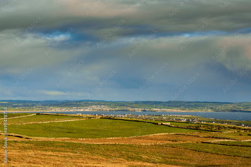Huge green fields in foreground and small town in the background by the ocean. Lahinch area, county Clare, Ireland. Cloudy sky. Agriculture land farming industry. Irish landscape.