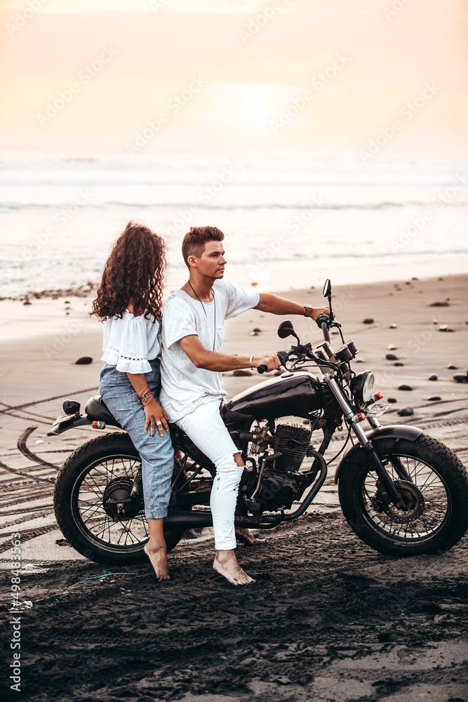 male and female couple in white outfit sitting on a motorcycle at a black sand beach in Bali Indonesia on a black vintage motorcycle during sunset
