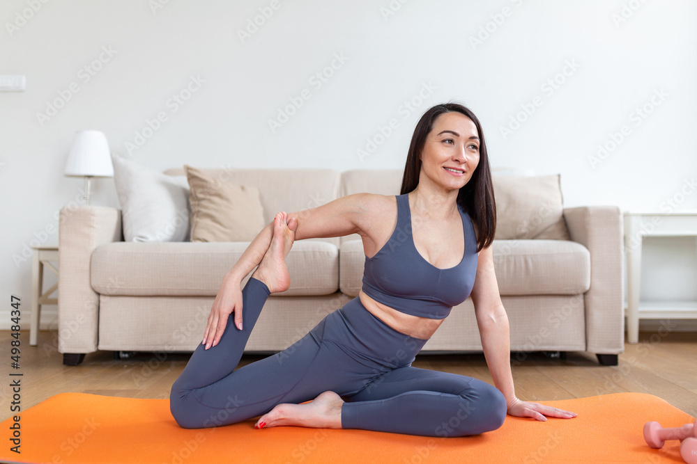 Young attractive smiling woman practicing yoga, sitting in Mermaid pose, working out, wearing sportswear, meditation session, indoor full length, home interior