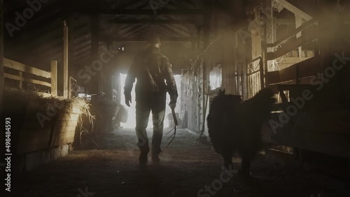 Cinematic shot of male farmer walking through the hay barn holding a pitchfork and calling out a dog. Sunlight breaks into the old farm house. An active shot of farmer walking. High quality 4k footage