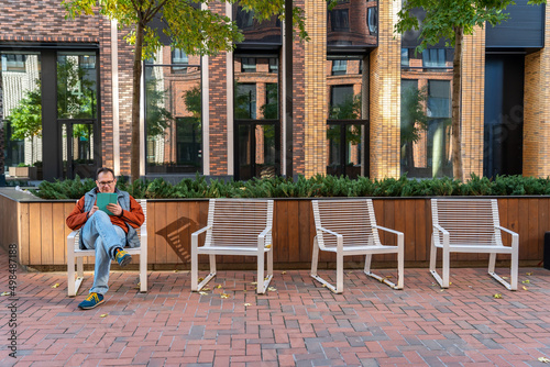 Fotobehang Millennial man with happy face checking phone sitting on one of four chairs in courtyard of modern  residential complex with landscaping