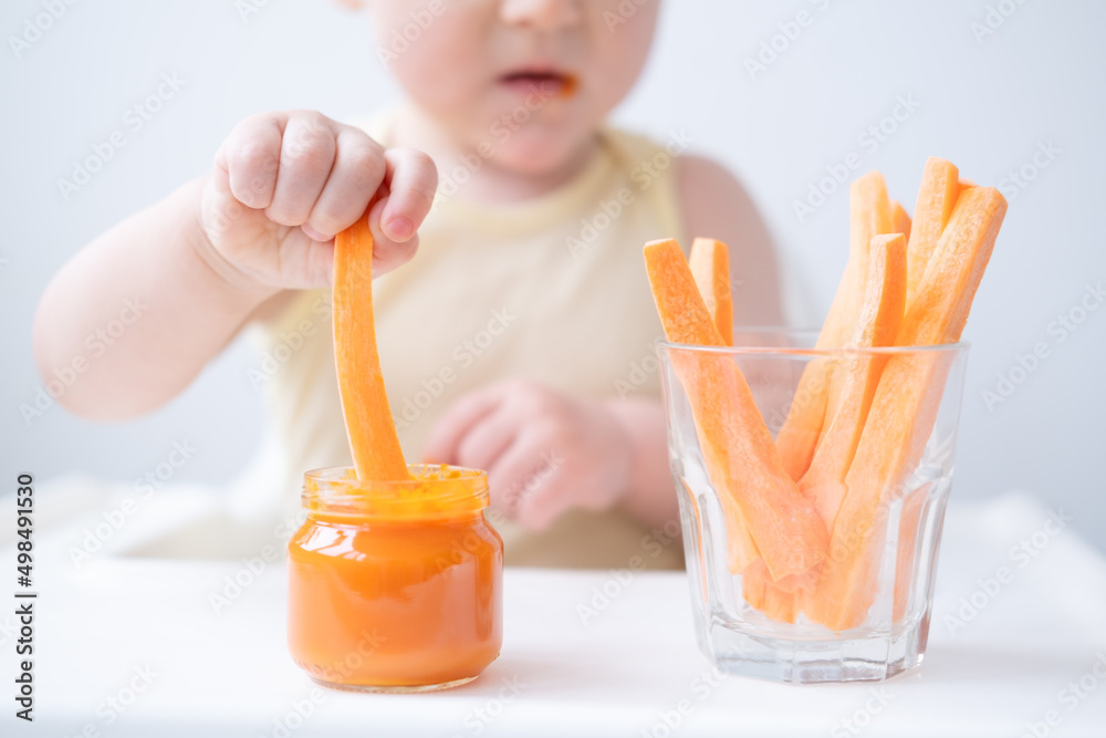 cute baby girl in yellow bodysuit sitting in childs chair eating carrot slices and vegetables purees cute baby girl in yellow bodysuit sitting in childs chair eating carrot slices and vegetables purees