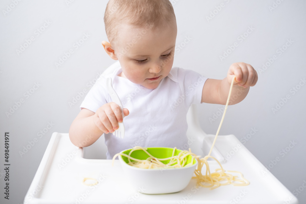 cute baby girl eating spaghetti pasta sitting in baby chair on white kitchen