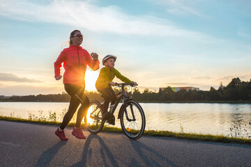 Happy mother and son go in sports outdoors. Boy rides bike in helmets, mom runs on sunny day. Silhouette family at sunset. Fresh air. Health care, authenticity, sense of balance and calmness