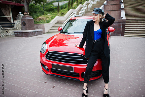 Portrait of beautiful blonde sexy fashion woman model in cap and in all black, blue corset, with bright makeup near red city car.