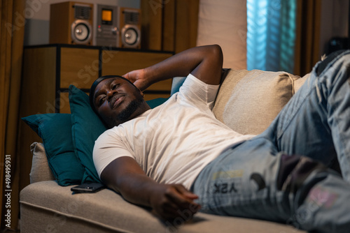 Man in white T-shirt and jeans lies on couch in front of the television at night. The student's room lit by warm color of the decorative lamps in living room. The men is relaxing while watching movie.