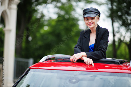 Portrait of beautiful blonde sexy fashion woman model in cap and in all black with bright makeup in red city car open sunroof.