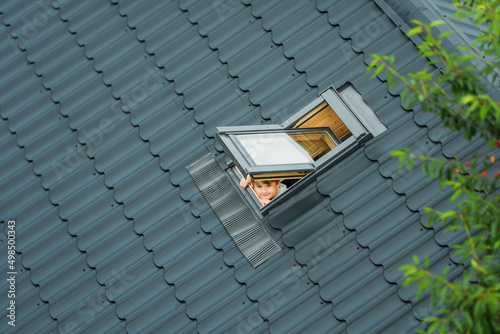 Εκτύπωση καμβά Little cheerful boy looks up into an open skylight window on roof of house covered with metallic gray tiles