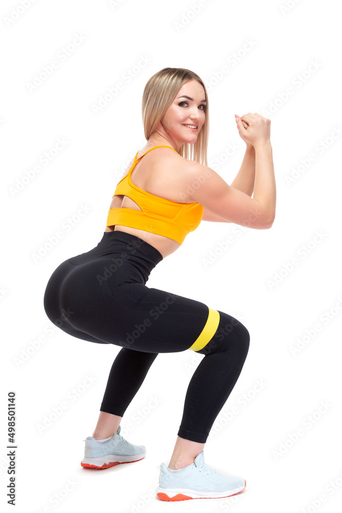 A beautiful, athletic, slim, smiling and cheerful woman in an orange top and black sweatpants performs squats with a fitness band. Lifestyle concept with sports and gym, healthy lifestyle. Isolated on A beautiful, athletic, slim, smiling and cheerful woman in an orange top and black sweatpants performs squats with a fitness band. Lifestyle concept with sports and gym, healthy lifestyle. Isolated on