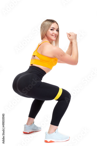 A beautiful, athletic, slim, smiling and cheerful woman in an orange top and black sweatpants performs squats with a fitness band. Lifestyle concept with sports and gym, healthy lifestyle. Isolated on