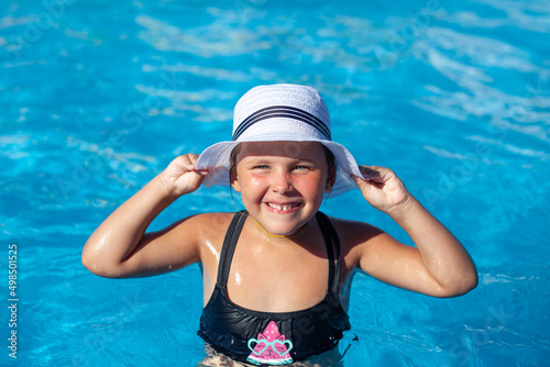 Little girl is relaxing in pool. Charming child in white straw hat decorated with ribbon is enjoying life and laughing with joy. 