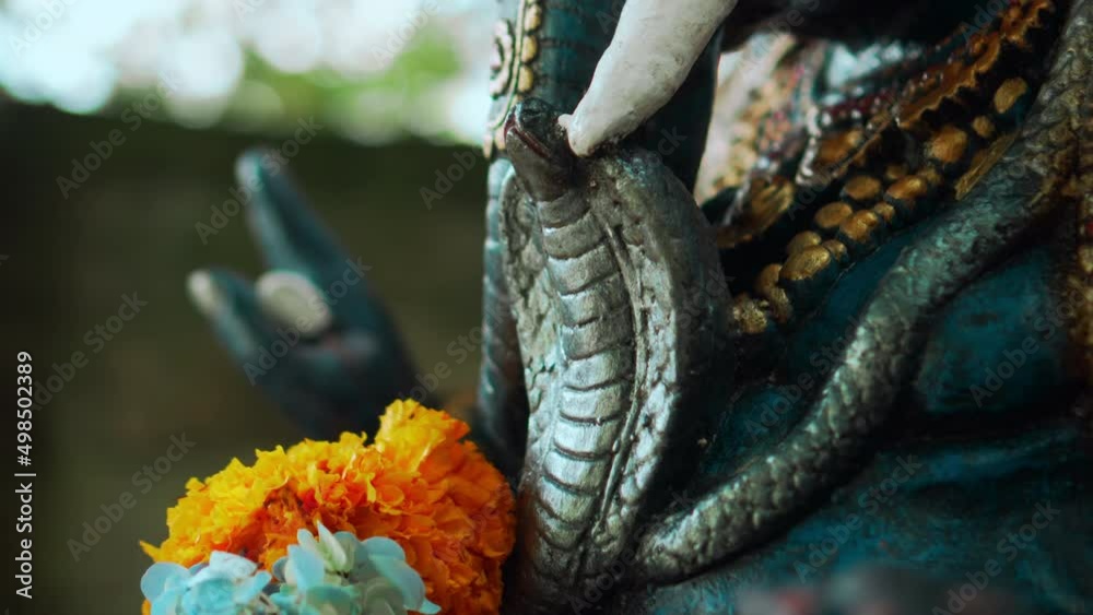 An old stone statue of Ganesha covered with moss with a wreath of yellow flowers on his neck standing on the street with green plants An old stone statue of Ganesha covered with moss with a wreath of yellow flowers on his neck standing on the street with green plants