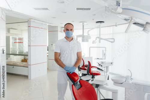 A young dentist in a mask stands near a red dental chair and smiles in modern white dentistry. Modern dentistry and prosthetics.