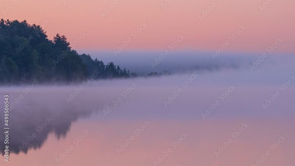 Dawn over the foggy lake. Beautiful dreamy view. Pink sky just before the sunrise and fog over water and trees with reflections on river bank.