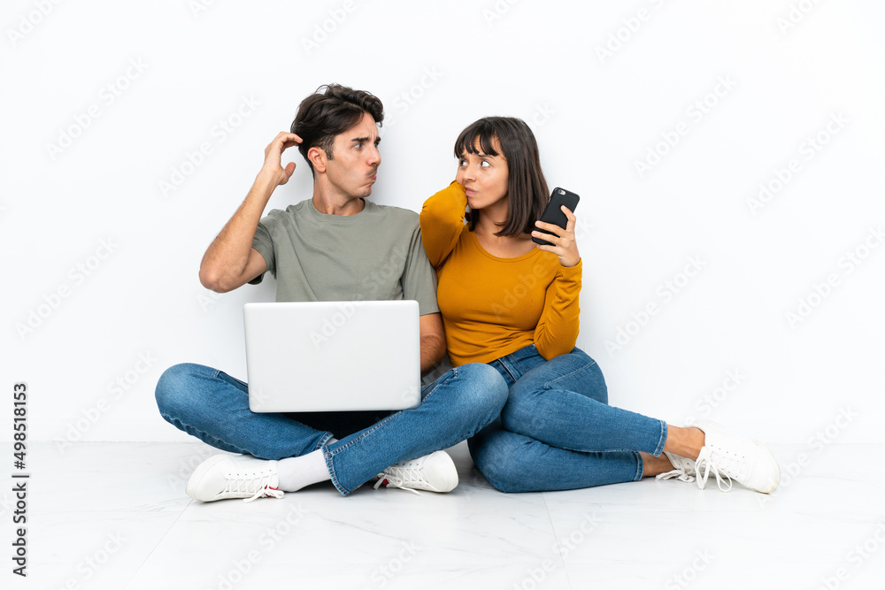 Young couple with a laptop and mobile sitting on the floor having doubts while scratching head Young couple with a laptop and mobile sitting on the floor having doubts while scratching head