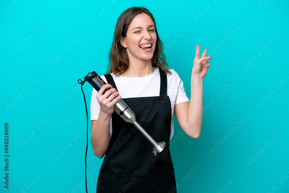 Young caucasian cooker woman using hand blender isolated on blue background smiling and showing victory sign Young caucasian cooker woman using hand blender isolated on blue background smiling and showing victory sign