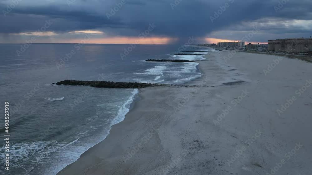 An aerial time lapse over a quiet beach in Arverne, NY as rain falls in the distance on a cloudy evening. The camera dolly out from a beautiful sunset, flying over the shores of the Atlantic Ocean.