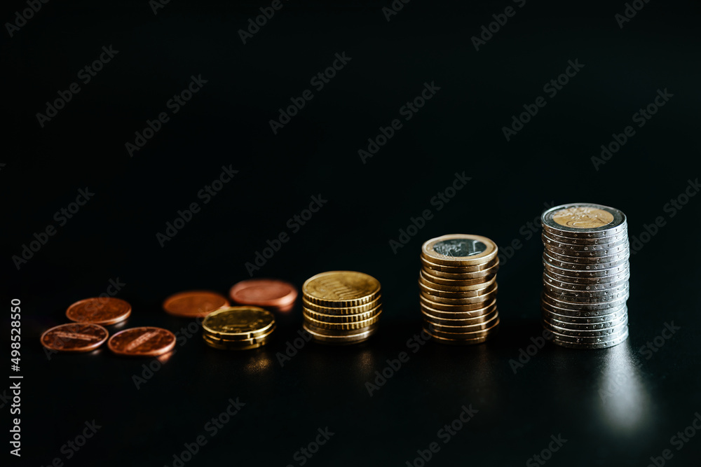 Euro coins stacked on each other with several pile of smaller coins on table isolated on black background, copy space. Earning, fund, future saving and money investment concept. Euro coins stacked on each other with several pile of smaller coins on table isolated on black background, copy space. Earning, fund, future saving and money investment concept.