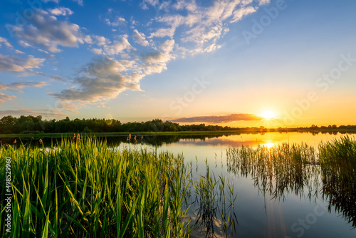 Fototapeta Naklejka Na Ścianę i Meble -  Amazing view at scenic landscape on a beautiful lake and colorful sunset with reflection on water surface among green reeds and glow on a background, spring season landscape