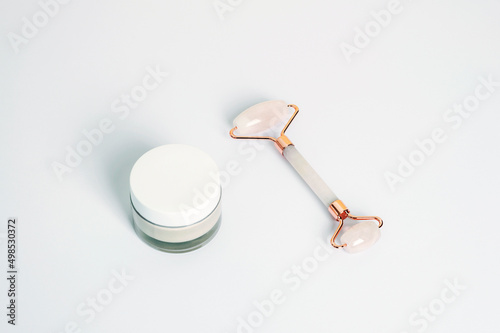 Cream in a jar and crystal rose quartz facial roller on white background, top view