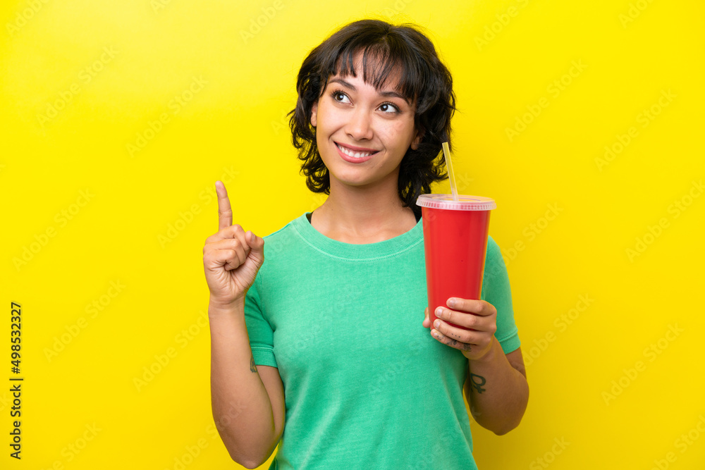 Young Argentinian woman holding a soda isolated on yellow background pointing up a great idea Young Argentinian woman holding a soda isolated on yellow background pointing up a great idea