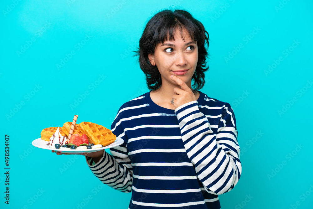 Young Argentinian woman holding waffles isolated on blue background having doubts while looking up