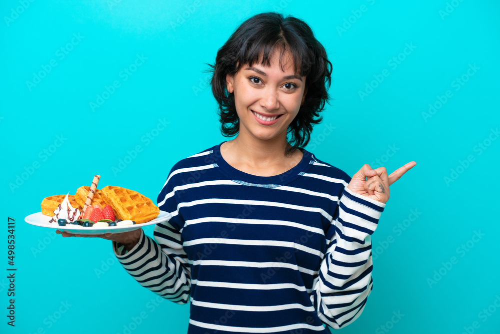 Young Argentinian woman holding waffles isolated on blue background pointing finger to the side Young Argentinian woman holding waffles isolated on blue background pointing finger to the side