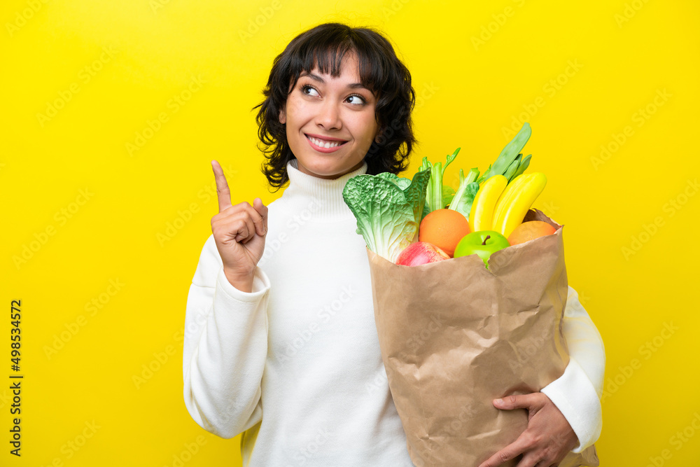 Young Argentinian woman holding a grocery shopping bag isolated on yellow background pointing up a great idea Young Argentinian woman holding a grocery shopping bag isolated on yellow background pointing up a great idea