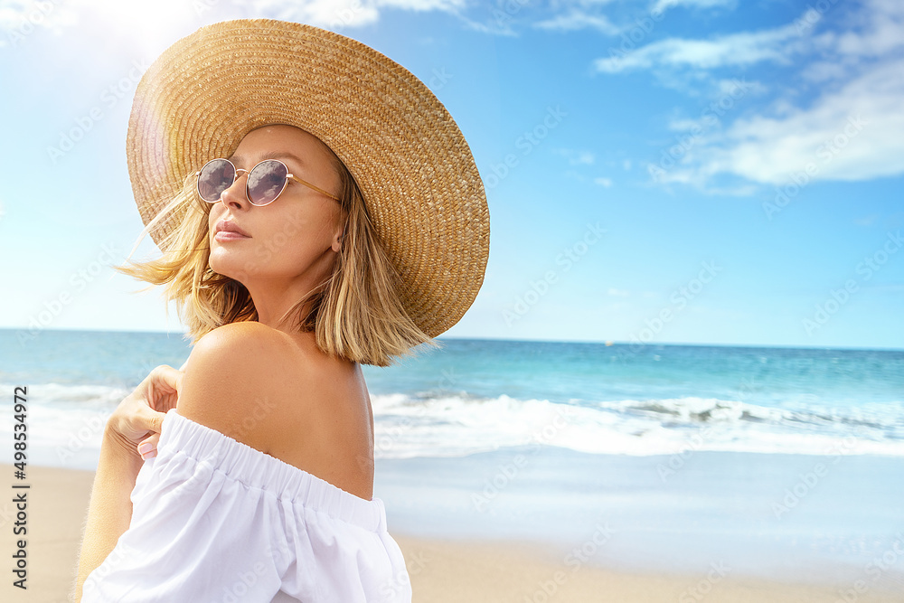 Portrait of blonde caucasian girl in summer straw hat and fashionable sunglasses walking, relaxing at the beach. Vacation vibe. Sunny day. Travel, tourism concept.