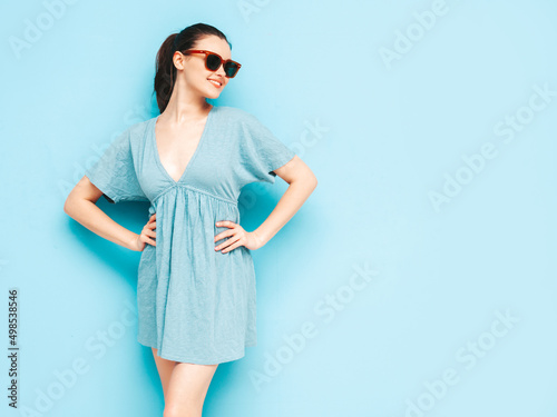 Portrait of young beautiful smiling female in trendy summer blue dress. Sexy carefree woman posing near blue wall in studio. Positive model having fun and going crazy. Cheerful and happy.In sunglasses