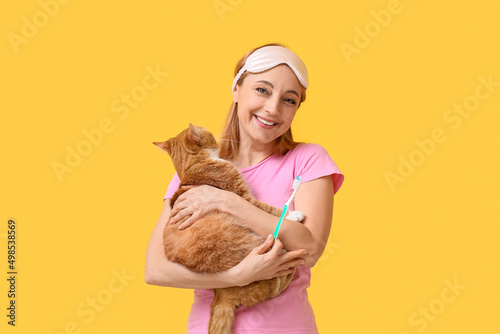 Beautiful mature woman with cute cat brushing teeth on yellow background