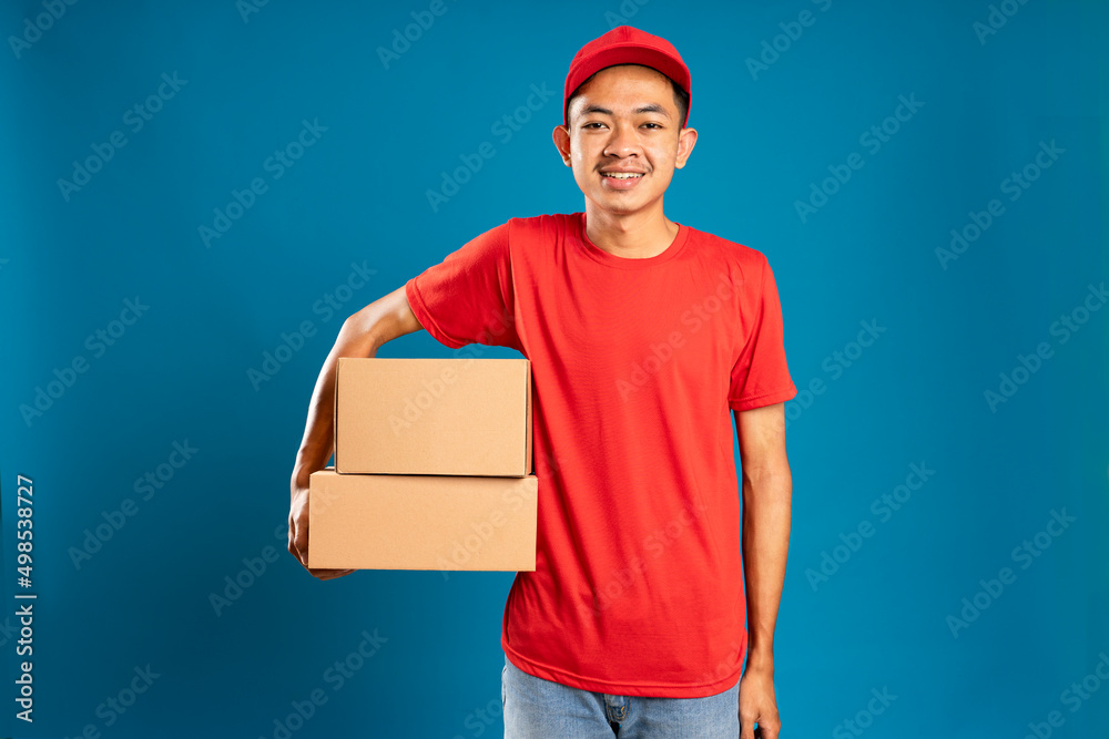 Happy young courier holding a cardboard box and smiling while standing against blue background Happy young courier holding a cardboard box and smiling while standing against blue background