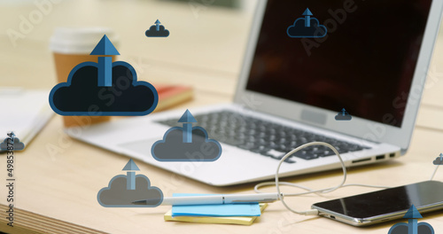 Image of clouds and arrows icons over smartphone and laptop on desk