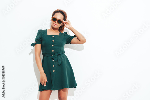 Beautiful black woman with afro curls hairstyle. Smiling model dressed in white summer green dress. Sexy carefree female posing near wall in studio. Tanned and cheerful. Isolated. In sunglasses