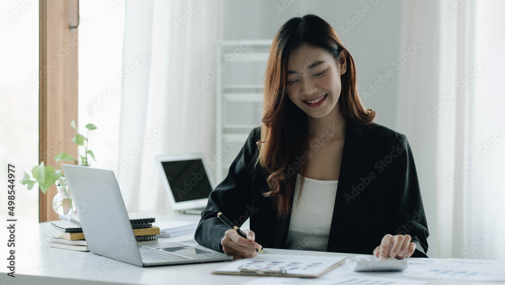 Beautiful Asian woman sitting in office using laptop and calculator. Happy business woman sitting at a desk in an office with a tablet computer. Beautiful Asian woman sitting in office using laptop and calculator. Happy business woman sitting at a desk in an office with a tablet computer.