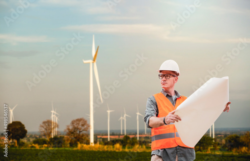 Handsome engineer wear protective helmets holding blueprint plan outdoors in work area with wind turbines in background.