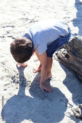 4-year-old boy dressed and barefoot playing with the sand on the beach.